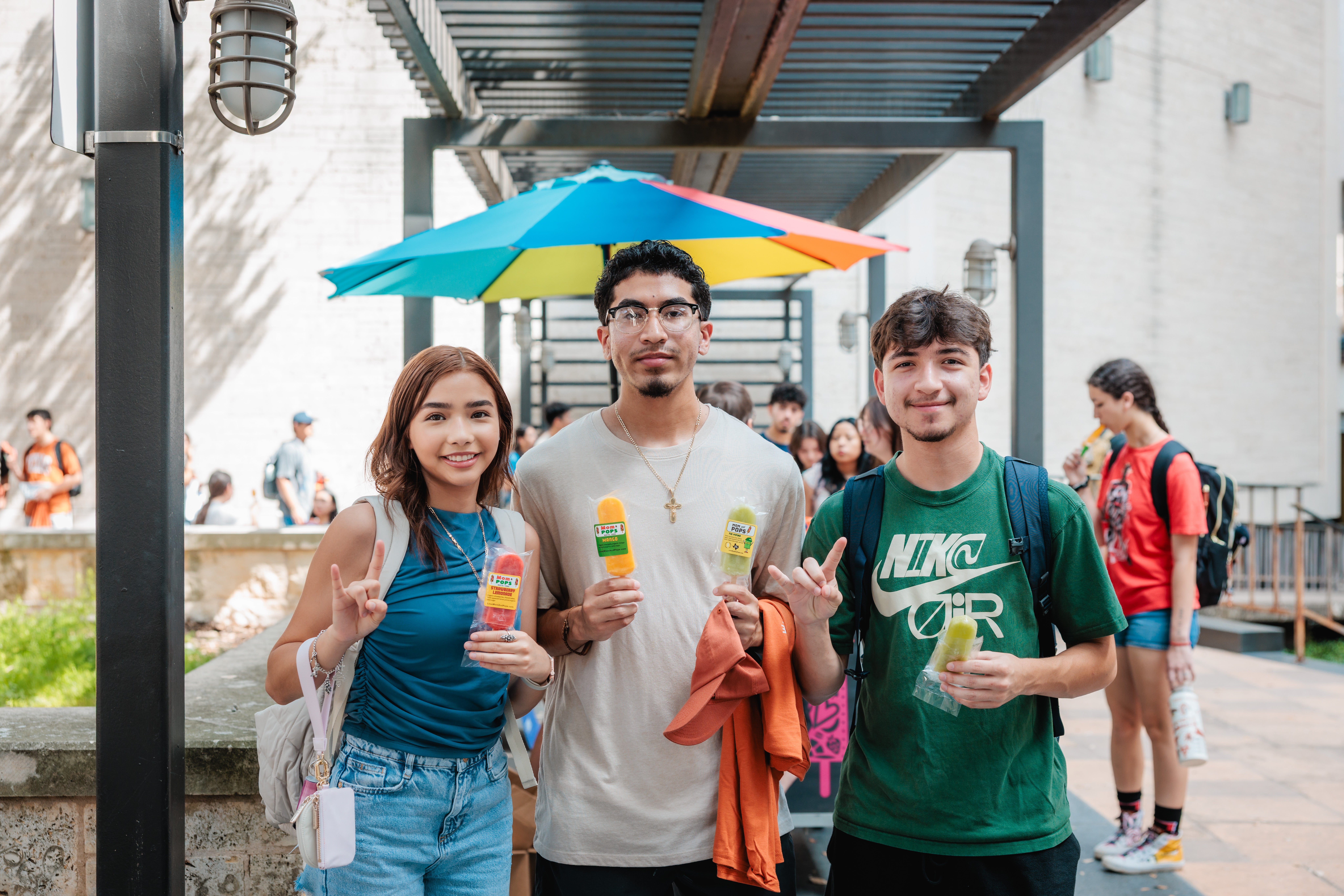 Three UT Austin students smiling and giving the Hook 'em Horns sign while holding fruit popsicles at an outdoor campus event, with a colorful umbrella and crowd of students visible in the background.